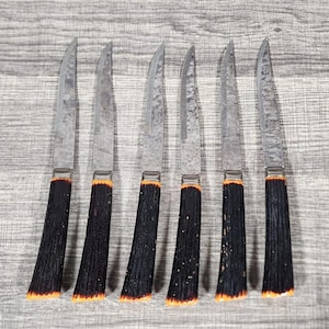 May include: A set of six steak knives featuring serrated, hammered-finish metal blades and dark, textured handles with orange-toned accents at the base. Each knife has a slender, pointed tip and a metallic bolster connecting the blade to the handle.