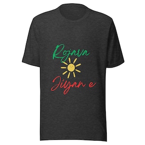 Könnte beinhalten: Dunkelgraues T-Shirt mit den Worten "Rojava Jiyan e" in grüner und roter Schrift und einer gelben Sonnengrafik in der Mitte. Das Shirt besteht aus einem weichen Material.