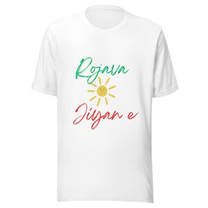 May include: White t-shirt with the words "Rojava Jiyan e" in green and red script, with a yellow sun graphic in between. The shirt is made of a soft material and has a crew neck.