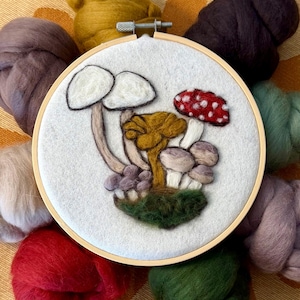 Op de afbeelding: Een vilten paddenstoelborduurwerk in een houten ring. De paddenstoelen zijn wit, rood, bruin en geel, met groen mos aan de basis. Rondom de ring liggen kleurrijke wolbundels in tinten rood, groen en paars.