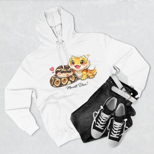 May include: A white hooded sweatshirt featuring a graphic illustration of a brown and black ball python and a yellow bearded dragon with the text Muzett Diaz. The hoodie is paired with black denim jeans and black and white canvas sneakers.