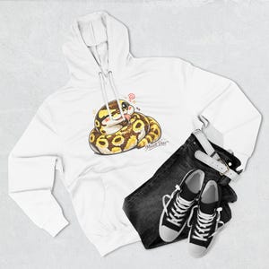 May include: A white hooded sweatshirt featuring a graphic of a coiled yellow and brown snake with a cartoon face and the text Muzett Diaz. The hoodie is paired with dark gray denim jeans and a pair of black and white high-top canvas sneakers.
