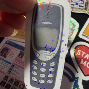 Op de afbeelding: Een gestanste sticker met een donkergrijze vintage Nokia mobiele telefoon met een numeriek toetsenbord en een scherm. De sticker heeft een holografische afwerking met verspreide kleurrijke glitterachtige vlekjes over het oppervlak.