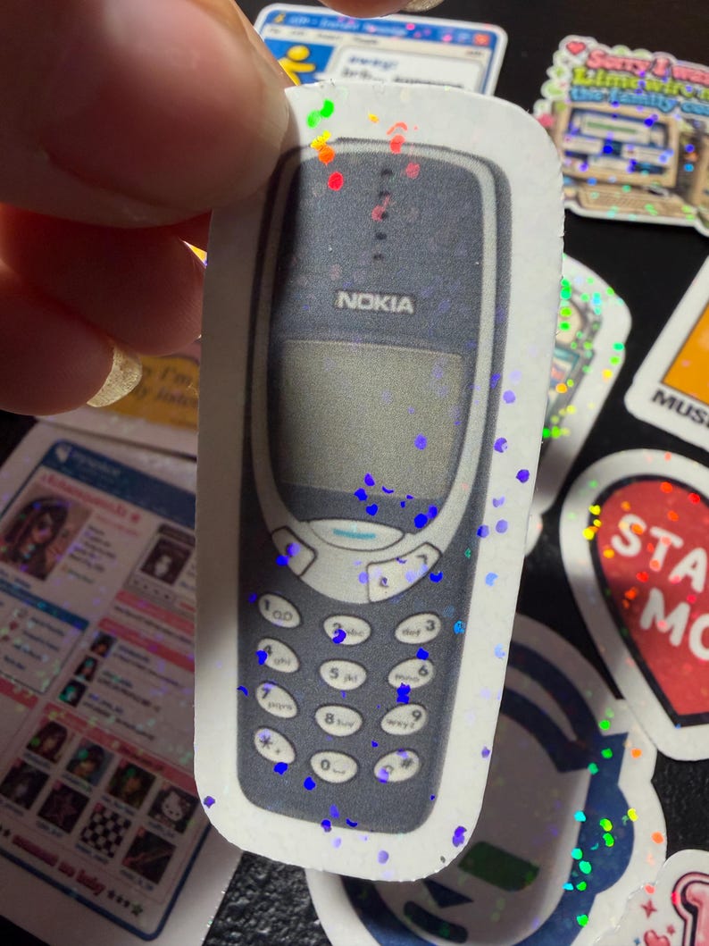Op de afbeelding: Een gestanste sticker met een donkergrijze vintage Nokia mobiele telefoon met een numeriek toetsenbord en een scherm. De sticker heeft een holografische afwerking met verspreide kleurrijke glitterachtige vlekjes over het oppervlak.