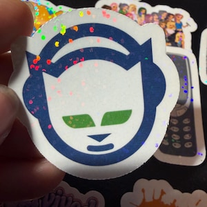 Op de afbeelding: Een ronde holografische sticker met een gestileerd donkerblauw kattenkop-icoon dat een koptelefoon draagt. De kat heeft felgroene ogen en een kleine donkerblauwe neus en mond. Het oppervlak van de sticker heeft een reflecterend glittereffect.