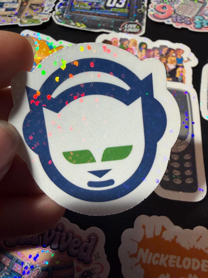 Op de afbeelding: Een ronde holografische sticker met een gestileerd donkerblauw kattenkop-icoon dat een koptelefoon draagt. De kat heeft felgroene ogen en een kleine donkerblauwe neus en mond. Het oppervlak van de sticker heeft een reflecterend glittereffect.