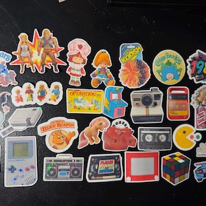 Op de afbeelding: Een verzameling kleurrijke vinylstickers met iconen uit de popcultuur van de jaren 80, waaronder een Game Boy, Rubik's Cube, Care Bears, He-Man, Strawberry Shortcake, een cassettebandje, een Polaroid-camera, Pac-Man en diverse retro speelgoed.