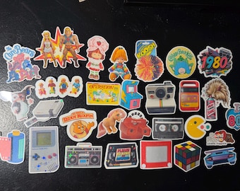 May include: A collection of colorful vinyl stickers featuring 1980s pop culture icons, including a Game Boy, Rubik's Cube, Care Bears, He-Man, Strawberry Shortcake, a cassette tape, a Polaroid camera, Pac-Man, and various retro toys and electronics.