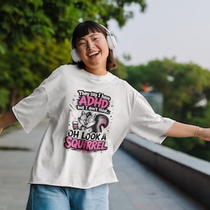 Op de afbeelding: Een wit T-shirt met korte mouwen en een grafische print van een eekhoorn die een kopje vasthoudt. De tekst luidt: They say I have ADHD but I don't think... OH LOOK A SQUIRREL. Het ontwerp bevat roze en zwarte kleuren met kleine sterren.