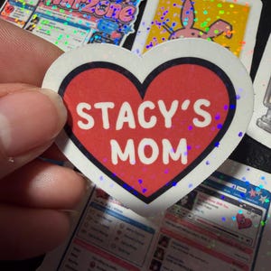 Op de afbeelding: Een hartvormige sticker met een rood midden en een dikke zwarte rand. De tekst STACY'S MOM is in dikke witte hoofdletters in het midden van het hart gedrukt. De sticker heeft een subtiele holografische afwerking die licht reflecteert.
