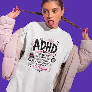 Op de afbeelding: Een wit t-shirt met een zwart en roze grafisch ontwerp met de tekst ADHD, YOUR SECRET'S SAFE WITH ME BECAUSE THERE'S A GOOD CHANCE I WASN'T LISTENING, vergezeld van een illustratie van een schedel met een roze strik en gekruiste botten.