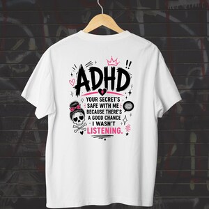 Op de afbeelding: Een wit t-shirt met een zwarte en roze afbeelding op de achterkant. Het ontwerp bevat de tekst "ADHD" en "YOUR SECRET'S SAFE WITH ME BECAUSE THERE'S A GOOD CHANCE I WASN'T LISTENING." Het toont ook een schedel met een roze strik en gekruiste botten.