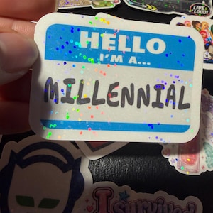 Op de afbeelding: Een rechthoekige sticker met een blauw-wit naamplaatjeontwerp met de tekst HELLO I'M A... MILLENNIAL in zwarte letters. De sticker heeft een holografische afwerking met verspreide kleurrijke glitterachtige stippen over het oppervlak.