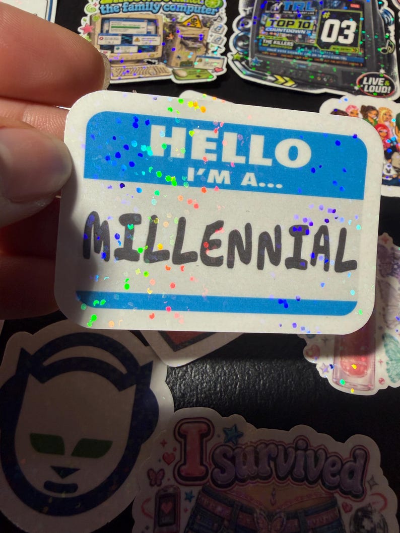 Op de afbeelding: Een rechthoekige sticker met een blauw-wit naamplaatjeontwerp met de tekst HELLO I'M A... MILLENNIAL in zwarte letters. De sticker heeft een holografische afwerking met verspreide kleurrijke glitterachtige stippen over het oppervlak.