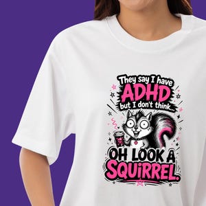 Op de afbeelding: Een wit t-shirt met een afbeelding van een eekhoorn die een koffiebeker vasthoudt. De tekst luidt: They say I have ADHD but I don't think... OH LOOK A SQUIRREL. Het ontwerp gebruikt zwarte en roze kleuren met sterren en een klein hartje.