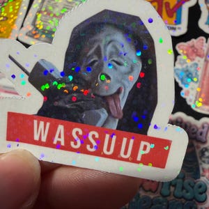 Op de afbeelding: Een holografische sticker met het Ghostface-personage uit de Scream-franchise, afgebeeld met de tong uit de mond terwijl het een vintage telefoon vasthoudt. Onder het personage staat een rode rechthoekige banner met de tekst WASSUUP.