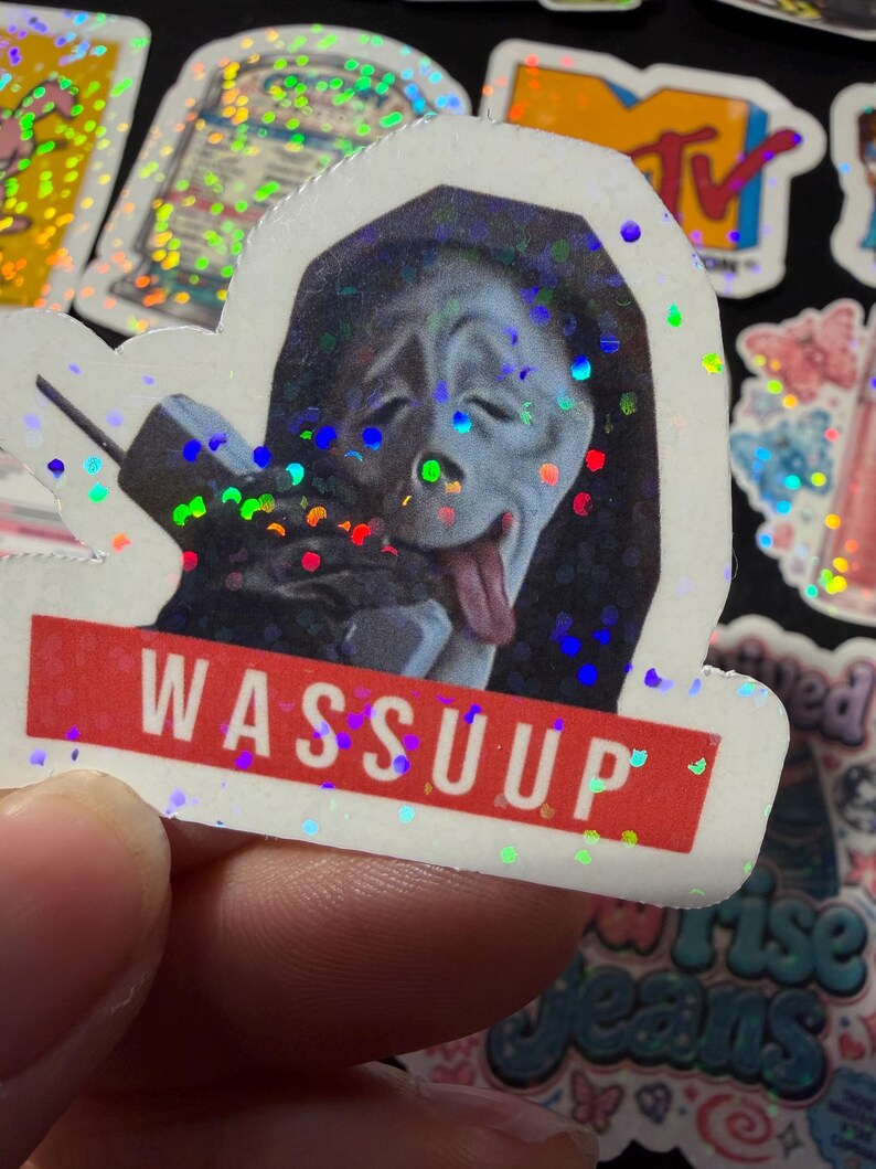 Op de afbeelding: Een holografische sticker met het Ghostface-personage uit de Scream-franchise, afgebeeld met de tong uit de mond terwijl het een vintage telefoon vasthoudt. Onder het personage staat een rode rechthoekige banner met de tekst WASSUUP.
