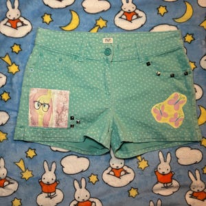 May include: Teal denim shorts with white polka dots, featuring a patch with a cartoon character and a butterfly design. The shorts have studded accents and a button closure. The shorts are on a blue patterned background.