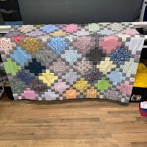 May include: A handmade quilt with a patchwork design featuring squares of various colors and patterns, including pink, blue, yellow, and gray. The quilt is displayed on a wooden surface, showcasing its intricate craftsmanship and vibrant hues.