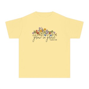 Könnte beinhalten: Ein hellgelbes T-Shirt mit einem Blumendesign und den Worten "grow in grace" in einer Schreibschrift. Die Blumenanordnung umfasst verschiedene bunte Blumen. Der Text "2 peter 3:18" steht darunter.