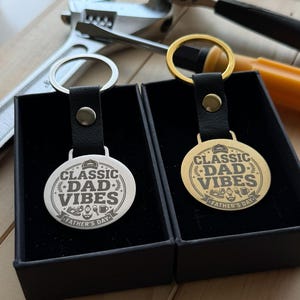 May include: Two circular keychains with black leather straps and metal rings, one silver and one gold. Each features an engraved design with the text "CLASSIC DAD VIBES" and "FATHER'S DAY" surrounded by small icons of a cap, shoes, watch, and mug.