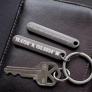 Peut inclure: Deux porte-clés en métal gris avec texte gravé : "A love that burns quiet and deep" et coordonnées "19.4756° N, 155.6054° W". Une clé et un porte-clés argenté sont attachés. Les porte-clés reposent sur une surface en cuir foncé.