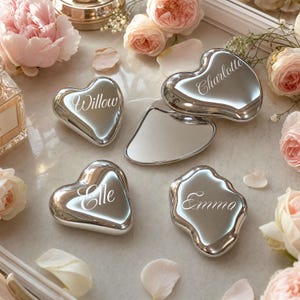 Peut inclure: Collection d'objets argentés en forme de cœur et de formes irrégulières, certains gravés de noms : "Willow", "Charlotte", "Elle" et "Emma". Les objets sont présentés avec des roses roses et des pétales.