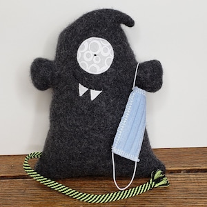 May include: A gray plush ghost toy with a white eye and teeth, wearing a light blue face mask. The ghost has a black and yellow striped cord attached to it.