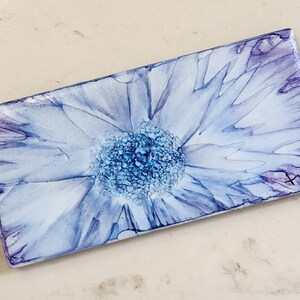 May include: A rectangular ceramic tile with a blue and purple watercolor flower design. The flower has a white center and blue petals.