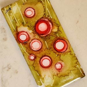 Puede incluir: Un azulejo rectangular con fondo verde y amarillo y círculos rojos y blancos. El azulejo tiene una superficie texturizada y un acabado brillante.