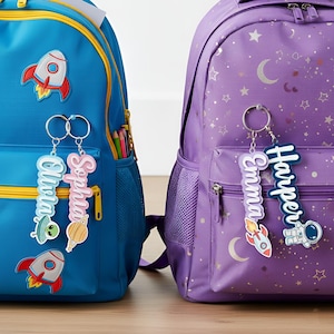 May include: Two backpacks, one blue and one purple, are shown. The blue backpack has rocket and alien patches, with keyrings that say "Olivia" and "Sophia". The purple backpack has star and moon patterns, with keyrings that say "Harper" and "Emma".