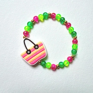 May include: A colorful beaded bracelet featuring a charm shaped like a striped beach bag. The bracelet is made of neon green, pink, and gold beads. The beach bag charm has pink, orange, and white stripes.