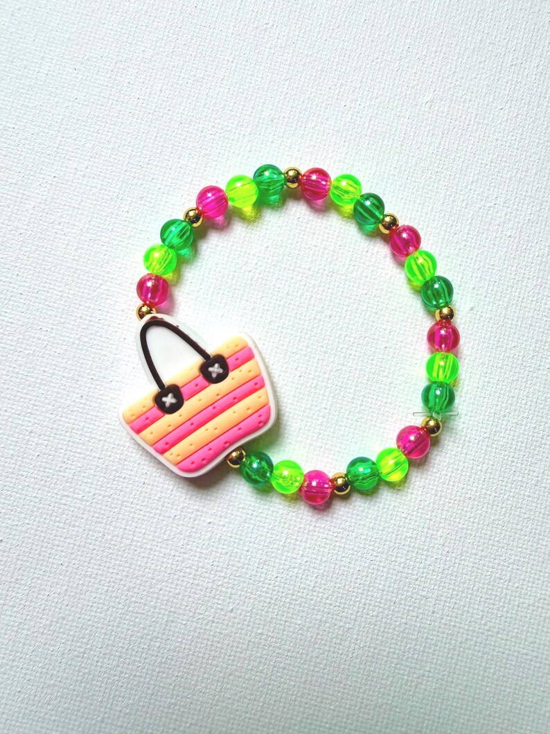 May include: A colorful beaded bracelet featuring a charm shaped like a striped beach bag. The bracelet is made of neon green, pink, and gold beads. The beach bag charm has pink, orange, and white stripes.