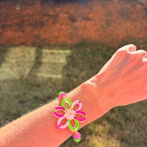 May include: A colorful flower bracelet with pink, green, and clear accents. The bracelet features a large flower design with a small dangling charm. The bracelet is worn on a wrist.