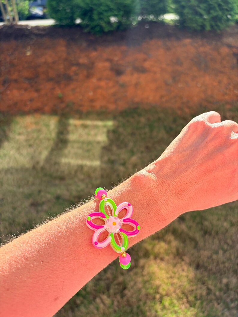 May include: A colorful flower bracelet with pink, green, and clear accents. The bracelet features a large flower design with a small dangling charm. The bracelet is worn on a wrist.