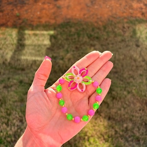 May include: A colorful bracelet featuring a flower-shaped charm and a beaded design. The bracelet has pink, green, and gold beads, with a matching flower charm in pink and green. The bracelet is a fun and playful accessory.