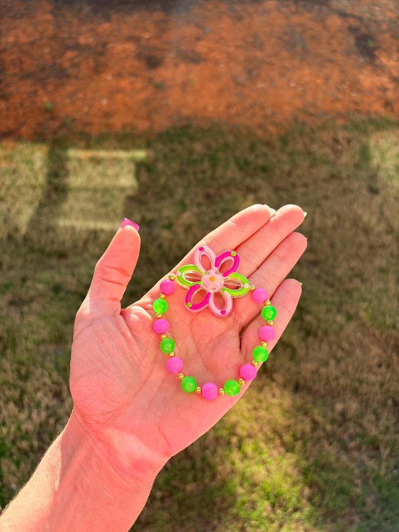 May include: A colorful bracelet featuring a flower-shaped charm and a beaded design. The bracelet has pink, green, and gold beads, with a matching flower charm in pink and green. The bracelet is a fun and playful accessory.
