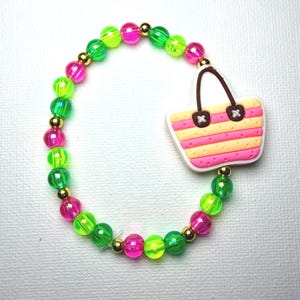 May include: A beaded bracelet with neon green, pink, and gold beads. The bracelet features a charm of a pink and yellow striped beach bag with brown handles. The bracelet is on a white background.