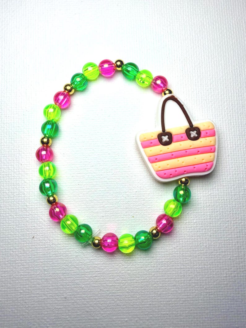 May include: A beaded bracelet with neon green, pink, and gold beads. The bracelet features a charm of a pink and yellow striped beach bag with brown handles. The bracelet is on a white background.