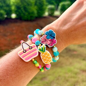 May include: Three beaded bracelets with tropical charms. One bracelet features a pink and yellow striped handbag charm. Another has a pineapple charm, and the third has a flower charm. The bracelets have pink, blue, and green beads.