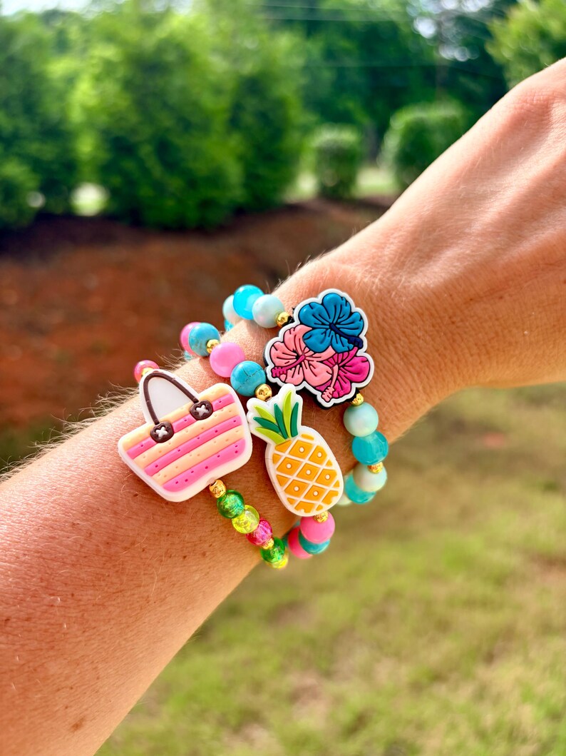 May include: Three beaded bracelets with tropical charms. One bracelet features a pink and yellow striped handbag charm. Another has a pineapple charm, and the third has a flower charm. The bracelets have pink, blue, and green beads.