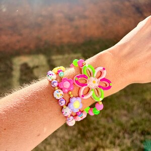 May include: A collection of colorful beaded bracelets featuring flower and round bead designs. The bracelets include pink, green, yellow, and purple beads. One bracelet has a large flower charm with pink and green accents.