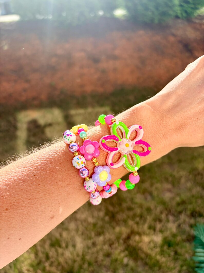 May include: A collection of colorful beaded bracelets featuring flower and round bead designs. The bracelets include pink, green, yellow, and purple beads. One bracelet has a large flower charm with pink and green accents.