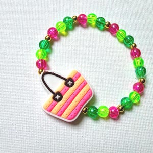 May include: A colorful beaded bracelet featuring a charm shaped like a handbag. The bracelet has green, pink, and gold beads. The handbag charm is pink, orange, and white with black details. A fun accessory.