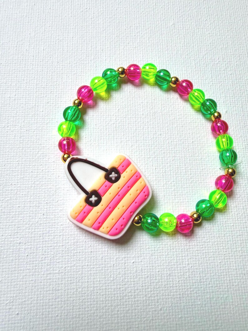 May include: A colorful beaded bracelet featuring a charm shaped like a handbag. The bracelet has green, pink, and gold beads. The handbag charm is pink, orange, and white with black details. A fun accessory.