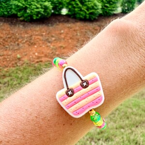 May include: A colorful bracelet featuring a charm shaped like a beach bag. The bag charm has pink and yellow stripes, brown accents, and a white handle. The bracelet includes rainbow-colored beads.