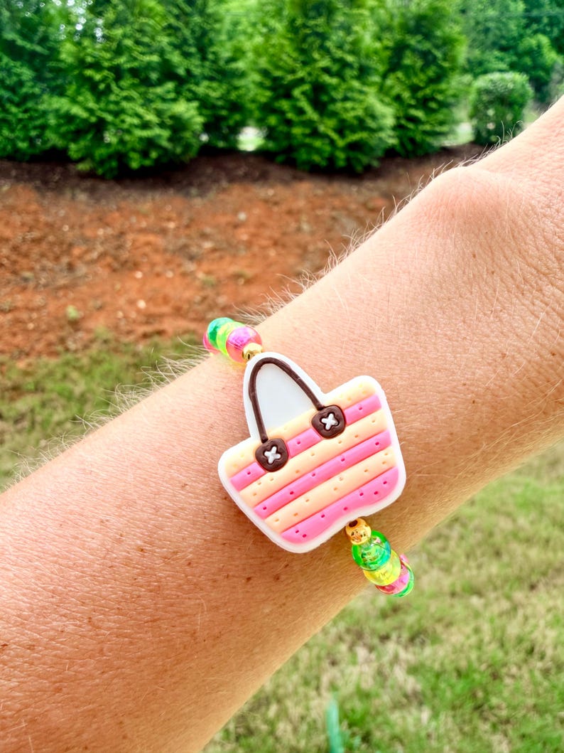 May include: A colorful bracelet featuring a charm shaped like a beach bag. The bag charm has pink and yellow stripes, brown accents, and a white handle. The bracelet includes rainbow-colored beads.