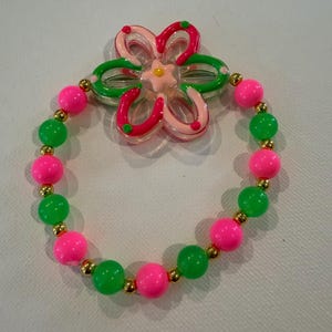 May include: A colorful bracelet with pink and green beads, interspersed with small gold-colored beads. A flower-shaped charm with pink, green, and clear accents sits above the bracelet. The bracelet is a fun, playful accessory.