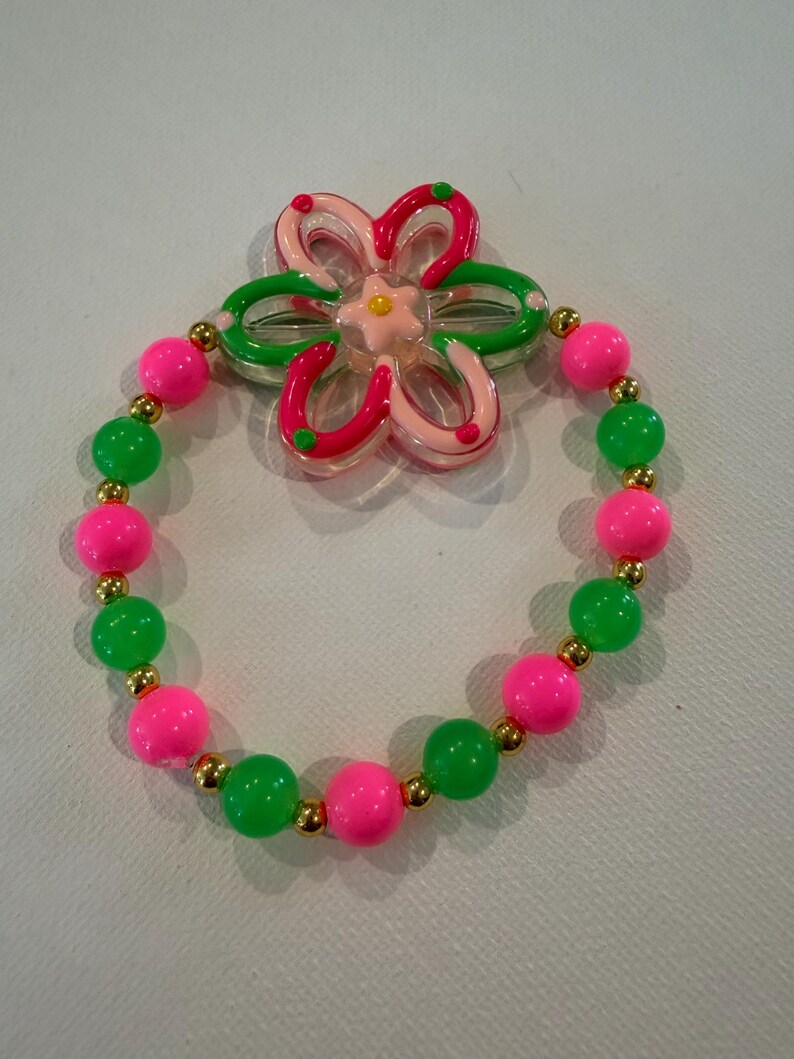 May include: A colorful bracelet with pink and green beads, interspersed with small gold-colored beads. A flower-shaped charm with pink, green, and clear accents sits above the bracelet. The bracelet is a fun, playful accessory.