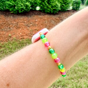 May include: A colorful beaded bracelet with pink, green, and yellow beads. The bracelet is accented with gold-colored spacers and a white and pink band. The bracelet is worn on a person's arm.